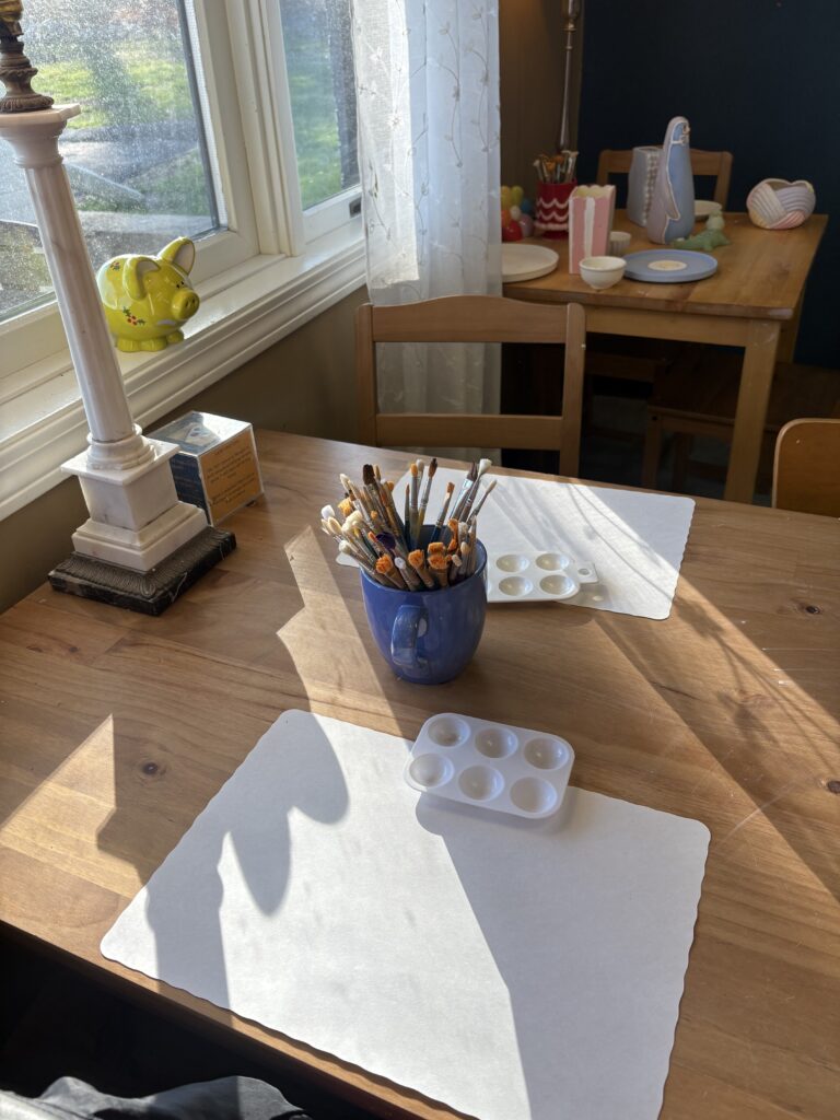 Two placemats and paint palettes are shown on a table bathed in sunlight. There is a cup of paint brushes in the middle of the table.