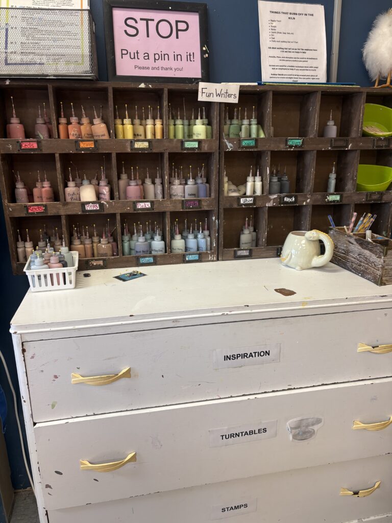 Colorful paint bottle writing tools are organized in a shelf by color. Sponges and dotting tools a for adding detail to the paint your own pottery pieces are shown