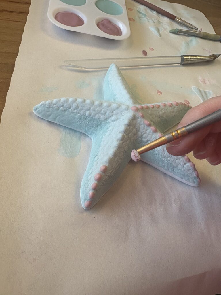 A dotter tool is being used to paint pink spots on the top of the light blue starfish piece