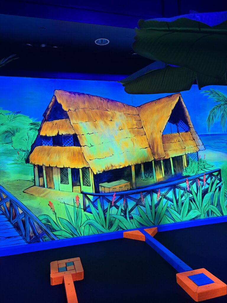 A mural of a hut with a glowing yellow roof is shown above a mini golf course