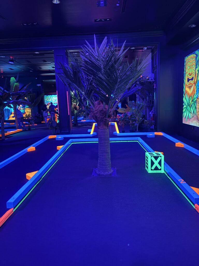 An indoor mini golf course is shown with a faux palm tree in the center