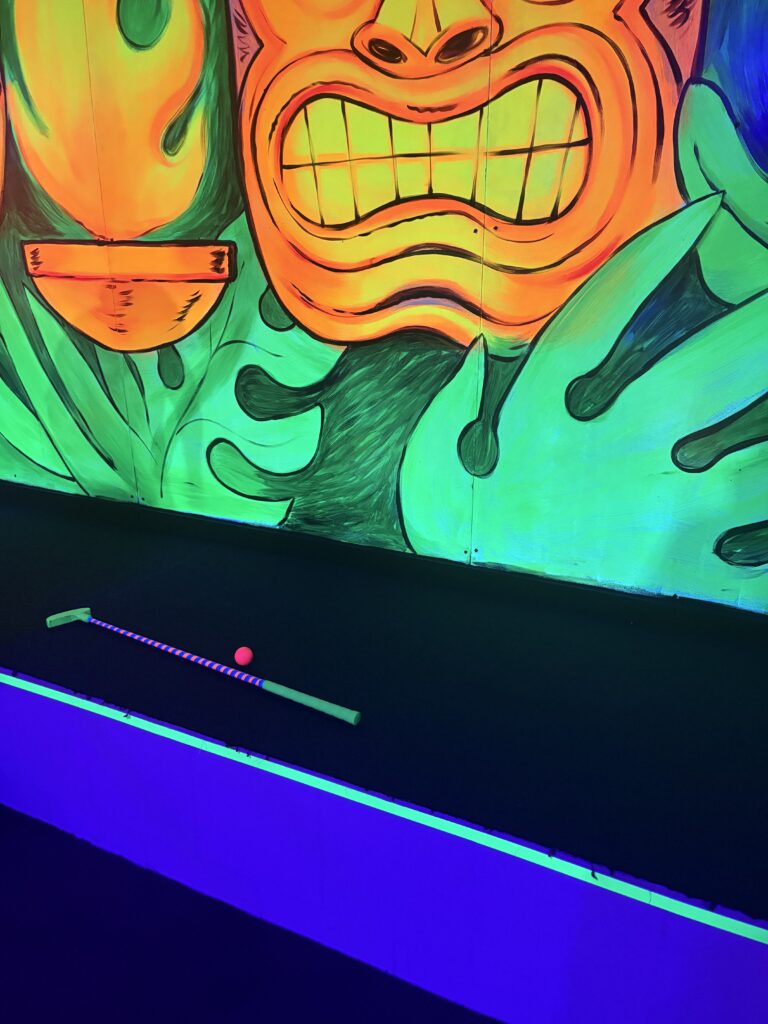 A ledge with a green putter and pink ball lying on it sits in front on a mural of a tiki figure.